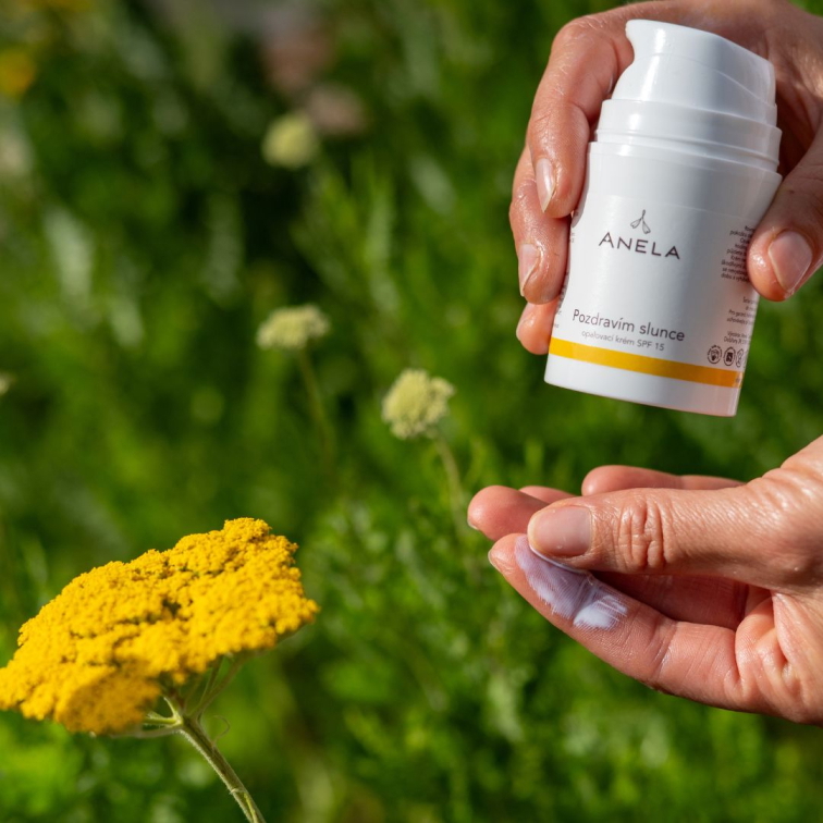 ruka roztírající výživný přírodní opalovací krém, v pozadí žlutá květina a lahvička s krémem