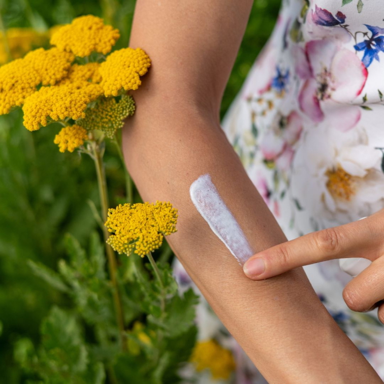 ruka roztírající výživný přírodní opalovací krém, v pozadí žlutá květina