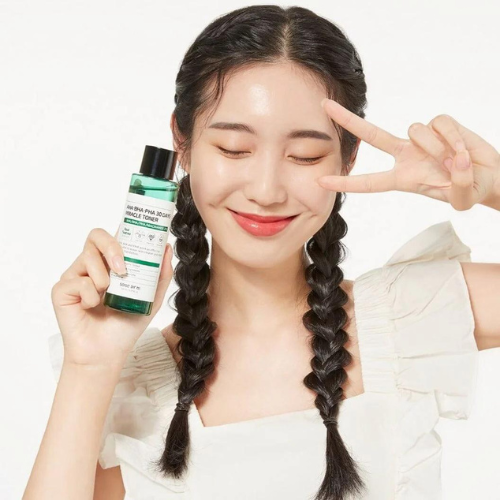 Krásná dívka se zářivou drží v ruce oblíbené korejské tonikum se 3 kyselinami - AHA·BHA·PHA 30 Days Miracle Toner a druhou rukou dvěmi vytváří V jako gesto vítězství.